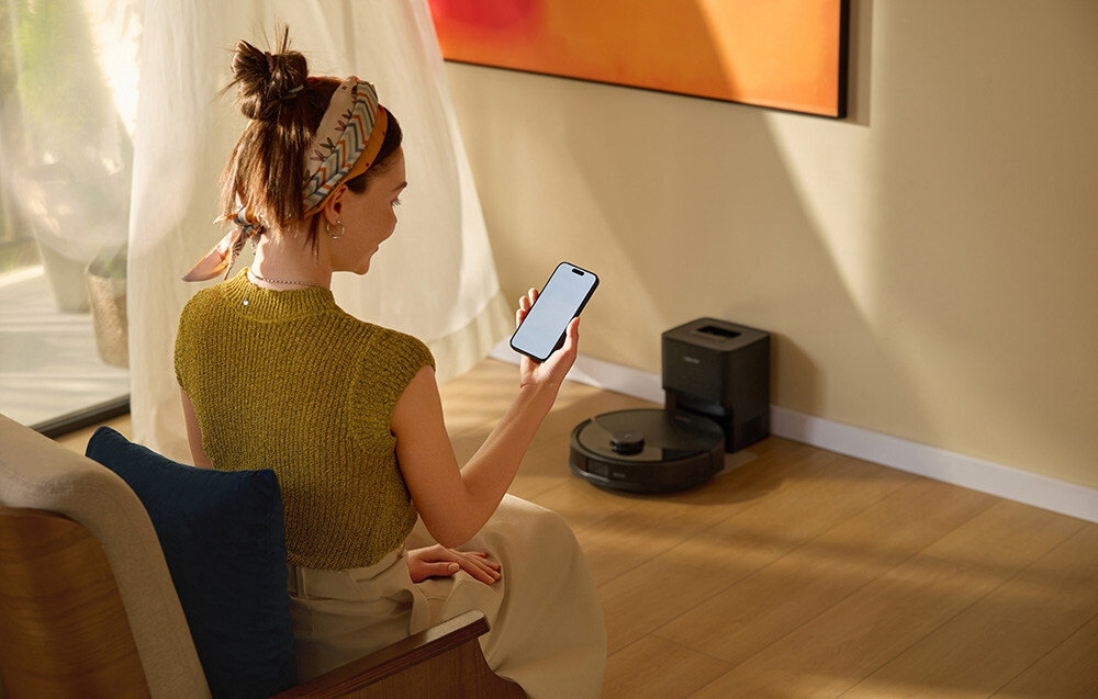 Osoba siedzi na sofie, trzymając w ręku smartfon, a na podłodze obok znajduje się czarny robot sprzątający z bazą ładującą. W tle widać zasłonę i fragment kolorowego obrazu na ścianie.