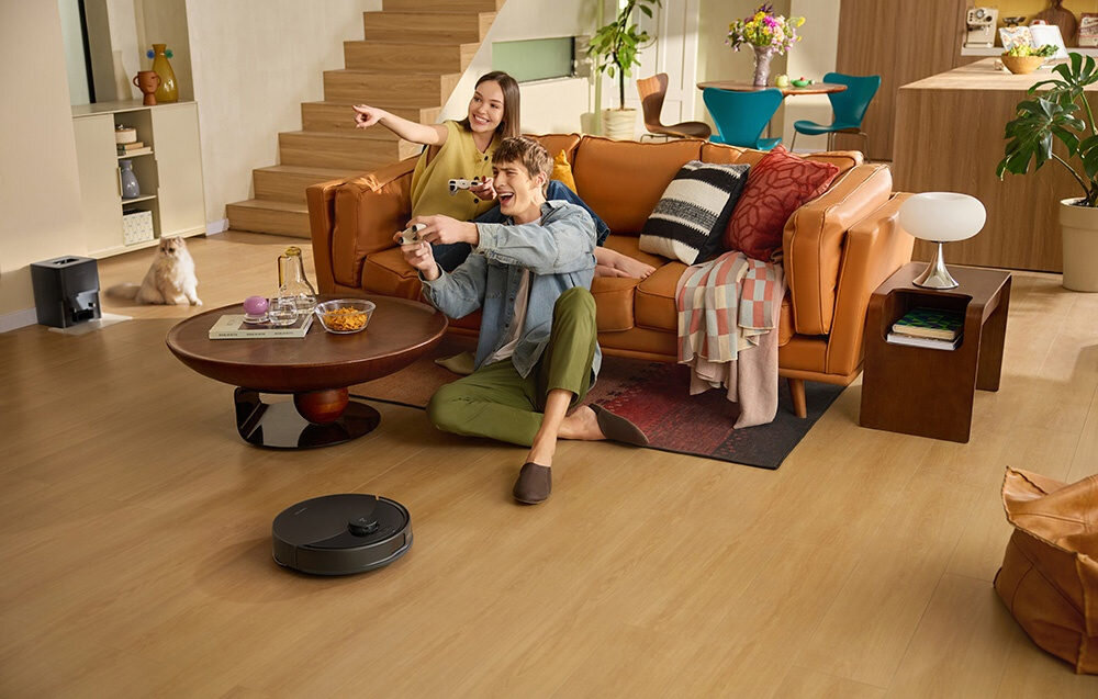 W salonie na drewnianej podłodze znajduje się czarny robot sprzątający. Na kanapie siedzą dwie osoby, a obok stolika stoi kot.