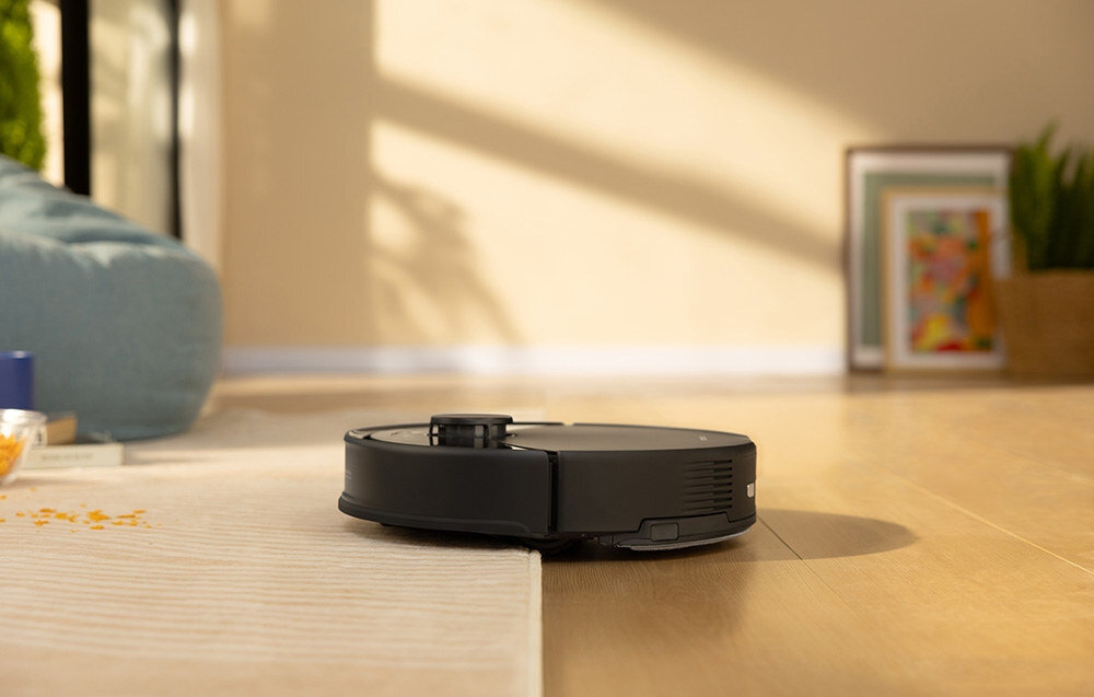 Czarny robot sprzątający porusza się po podłodze, częściowo na dywanie. W tle widać rozmyty obraz wnętrza z rośliną i obrazem na ścianie.