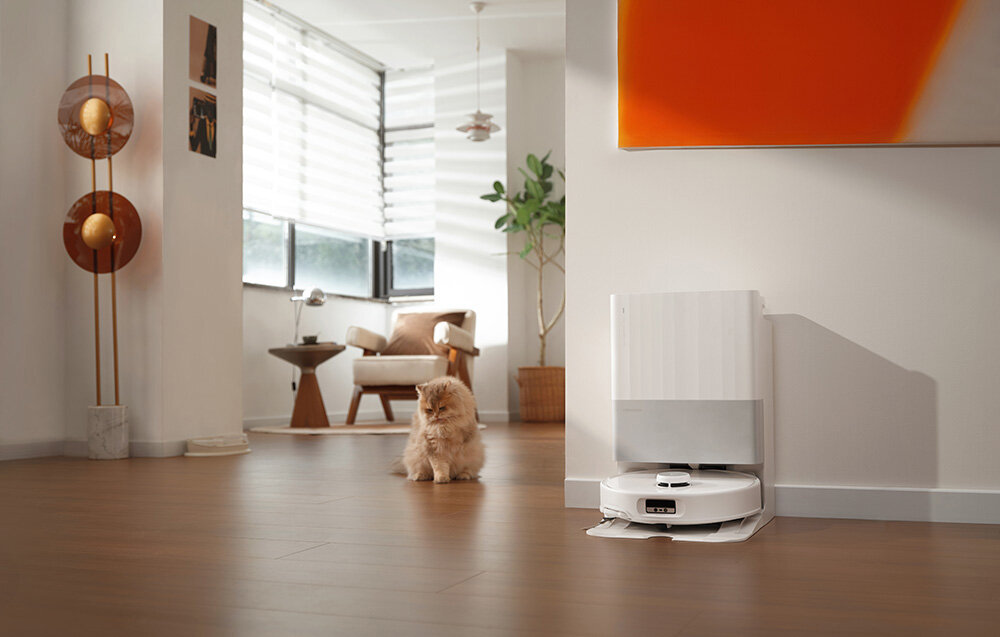 W jasnym wnętrzu na podłodze znajduje się biały robot sprzątający przy stacji dokującej. W tle widać fotel, stolik oraz roślinę, a na podłodze siedzi kot.