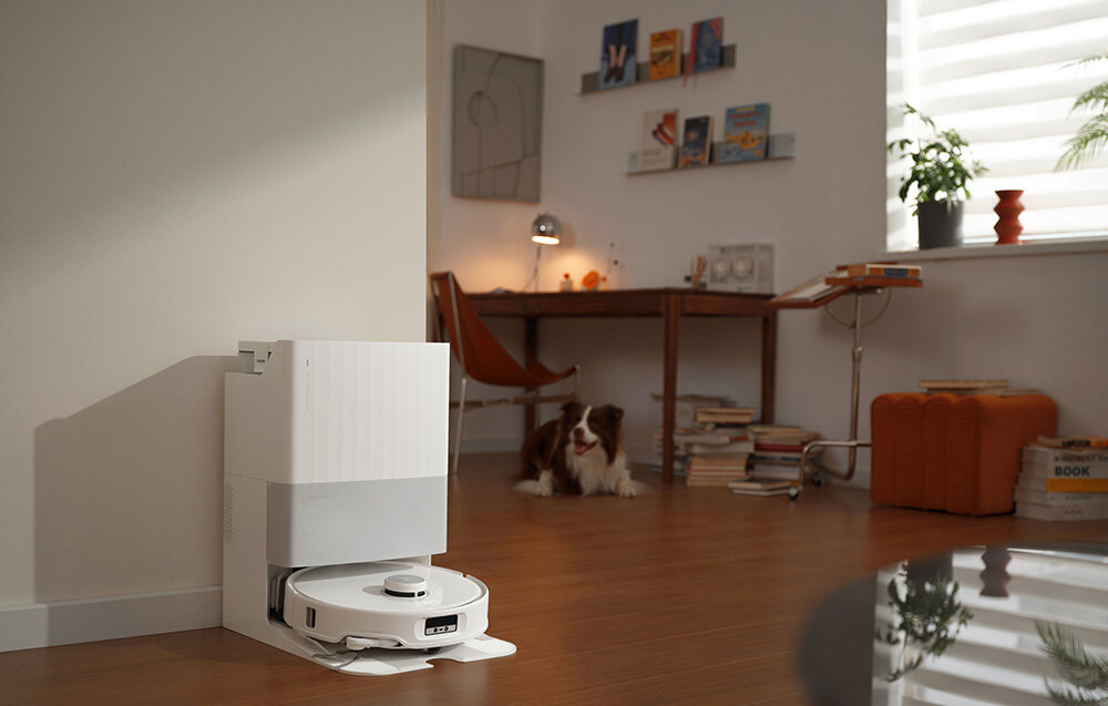 Na podłodze w pomieszczeniu znajduje się biały robot sprzątający w stacji dokującej. W tle widać biurko z lampką, książki oraz psa leżącego na podłodze.