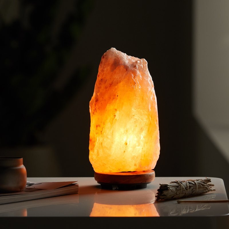 Lampa stołowa z naturalnego kamienia solnego emituje ciepłe, pomarańczowe światło. Stoi na drewnianej podstawie, obok niej leży zwinięta wiązka ziół i kilka książek.