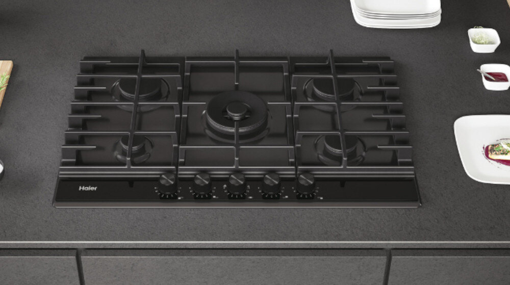 Czarna płyta gazowa z pięcioma palnikami i pokrętłami do regulacji. Widoczne logo \'Haier\' na przednim panelu.