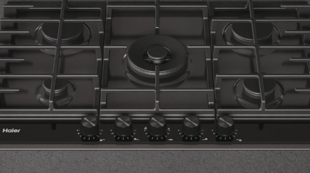 Na obrazie widoczna jest płyta gazowa z pięcioma palnikami i pokrętłami do regulacji. Logo \'Haier\' znajduje się w lewym dolnym rogu płyty.
