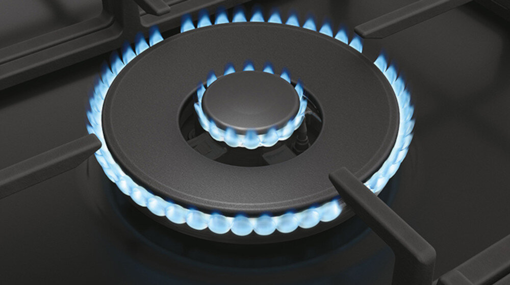 Obraz przedstawia palnik gazowy z niebieskim płomieniem na czarnej płycie kuchennej. Widać metalowe ruszty wokół palnika.