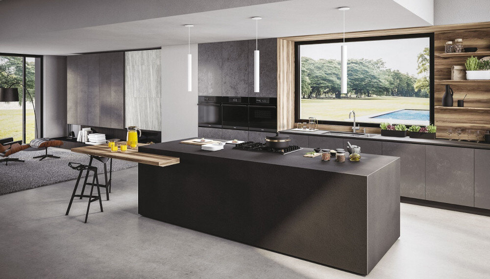 Nowoczesna kuchnia z dużą wyspą, na której znajduje się płyta gazowa i różne akcesoria kuchenne. W tle widoczne są duże okna z widokiem na zieleń oraz półki z dekoracjami.