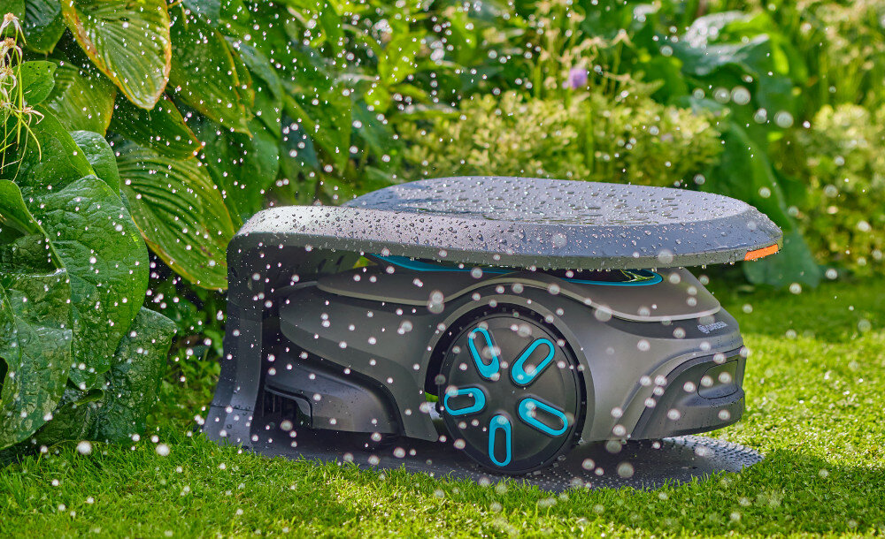 Na trawie znajduje się garaż dla robota koszącego, pokryty kroplami wody. W tle widoczne są zielone liście roślin.