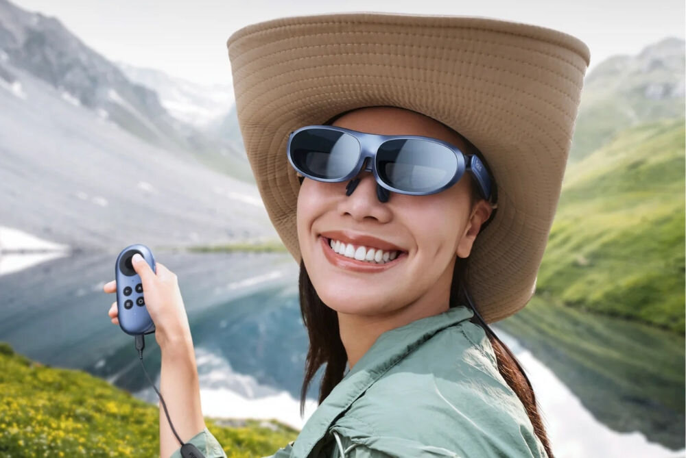 Osoba w kapeluszu nosi okulary i trzyma w ręku urządzenie z przyciskami. W tle widać górski krajobraz z jeziorem.