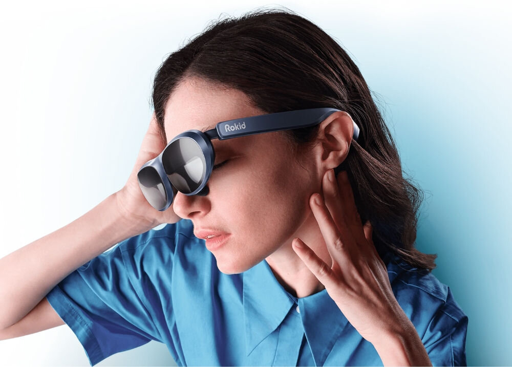 Osoba nosi niebieskie okulary z napisem \'Rokid\' na zauszniku. Tło jest jasnoniebieskie, a osoba ubrana jest w niebieską koszulę.