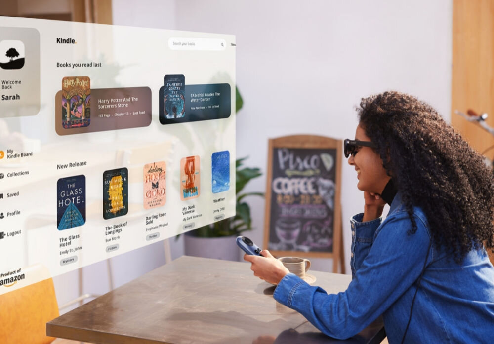 Osoba siedzi przy stole, nosząc okulary i trzymając urządzenie w ręku, patrzy na wyświetlany interfejs Kindle z listą książek. W tle widoczna jest tablica z napisem \'Please order coffee here\'.
