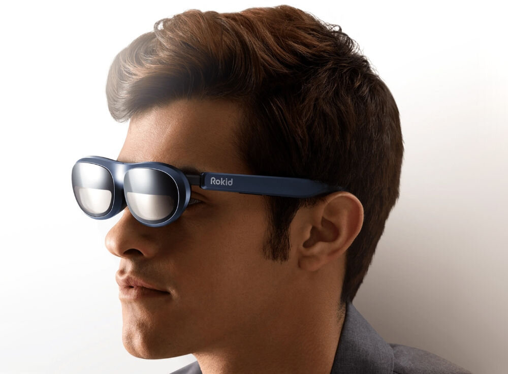 Osoba nosi niebieskie okulary z napisem \'Rokid\' na zauszniku. Tło jest jasne i jednolite.