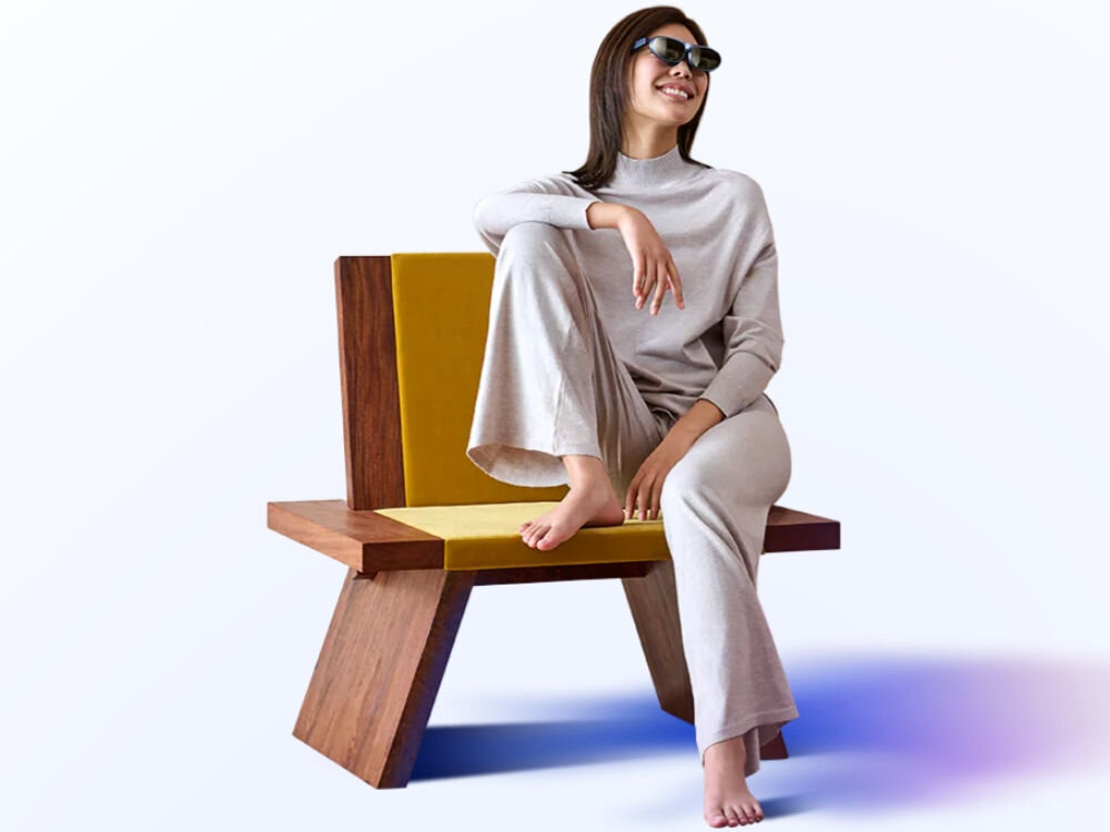 Osoba siedzi na nowoczesnym krześle z drewnianymi elementami i żółtym oparciem, ubrana w jasny strój i okulary przeciwsłoneczne. Tło jest jednolite i jasne.
