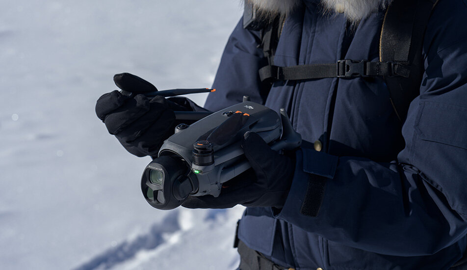 Osoba w ciemnej kurtce trzyma drona z dużą kamerą i kontrolerem. W tle widać śnieg.