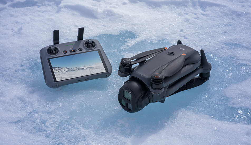 Na śniegu leży złożony dron oraz kontroler z ekranem wyświetlającym zimowy krajobraz. Dron ma ciemny kolor i kompaktową konstrukcję.