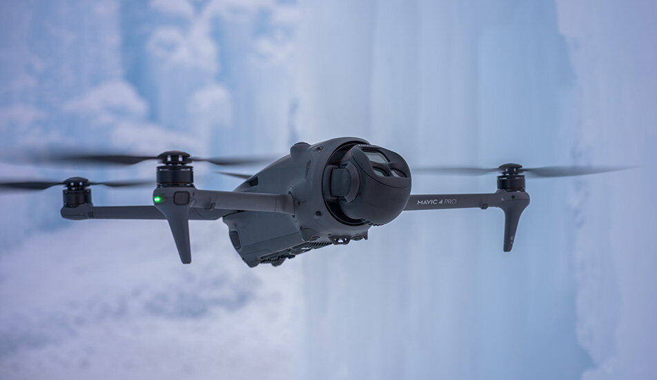 Dron z napisem \'MAVIC 4 PRO\' unosi się w powietrzu na tle lodowego krajobrazu. Widoczne są cztery śmigła i kamera z przodu urządzenia.