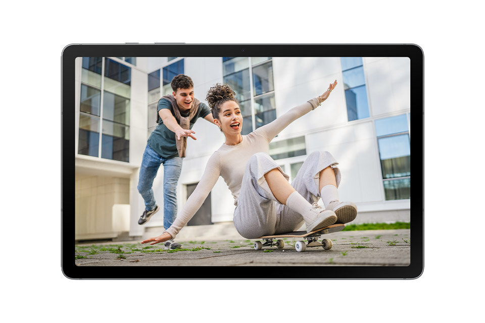 Tablet wyświetla obraz dwóch osób bawiących się na deskorolce przed nowoczesnym budynkiem. Ekran urządzenia jest włączony, a jego ramka jest czarna.
