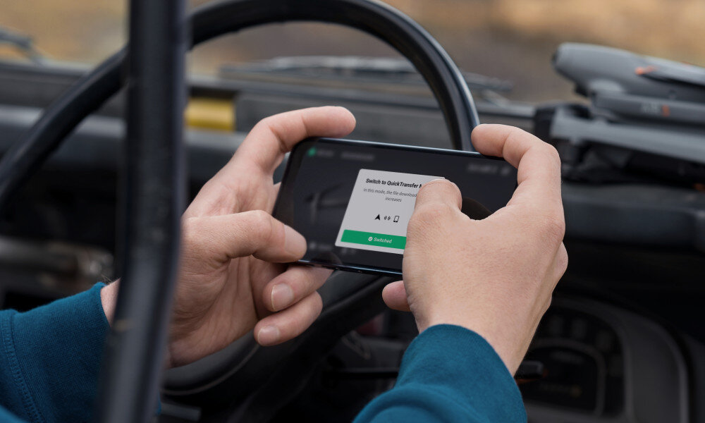 Osoba trzyma smartfon z wyświetlonym komunikatem \'Switch to QuickTransfer?\'. W tle widać wnętrze pojazdu i drona na desce rozdzielczej.