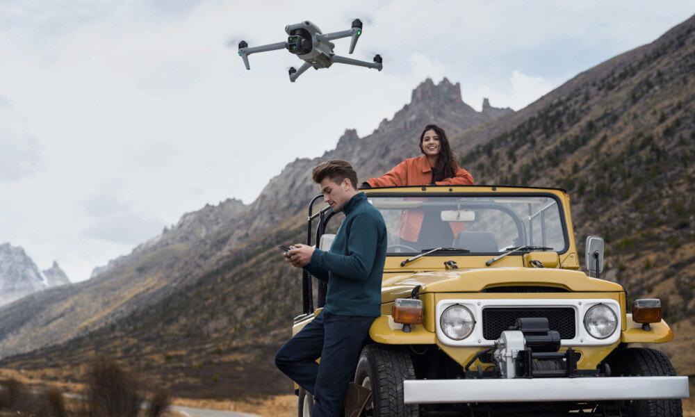 Dron unosi się w powietrzu nad żółtym samochodem terenowym, na którym siedzi kobieta, a obok stoi mężczyzna z kontrolerem. W tle widoczne są góry i pochmurne niebo.