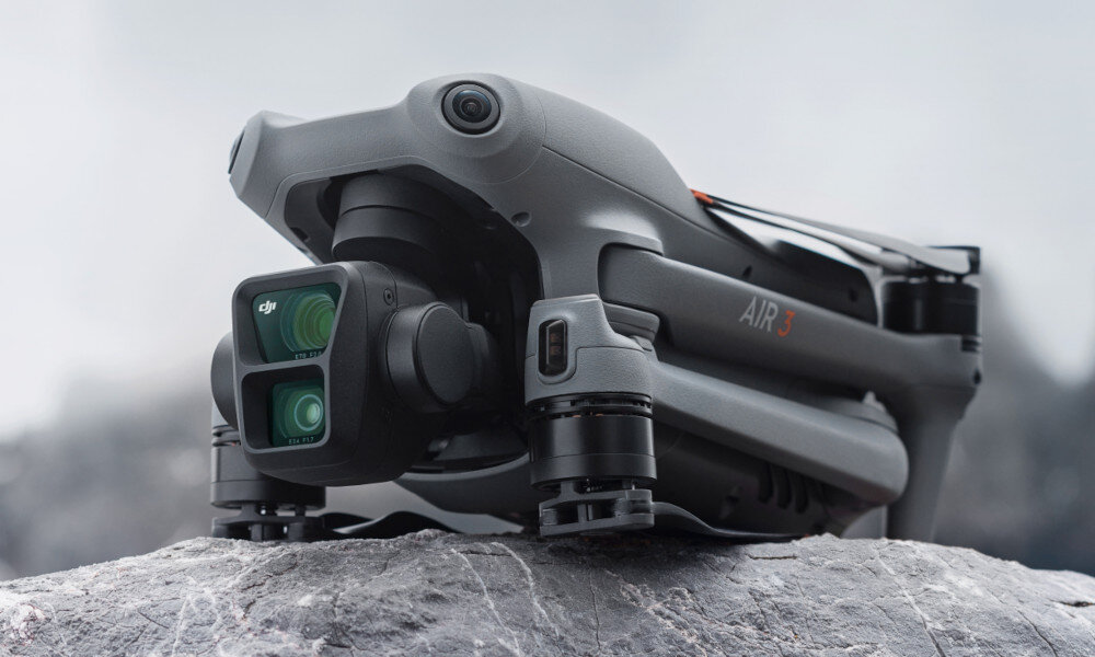 Na obrazie widoczny jest szary dron z napisem \'AIR 3\', umieszczony na kamieniu. Dron posiada podwójną kamerę z logo DJI.