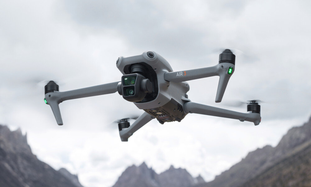 Dron z czterema wirnikami unosi się w powietrzu na tle górskiego krajobrazu. Na obudowie widoczny jest napis \'AIR 3\'.