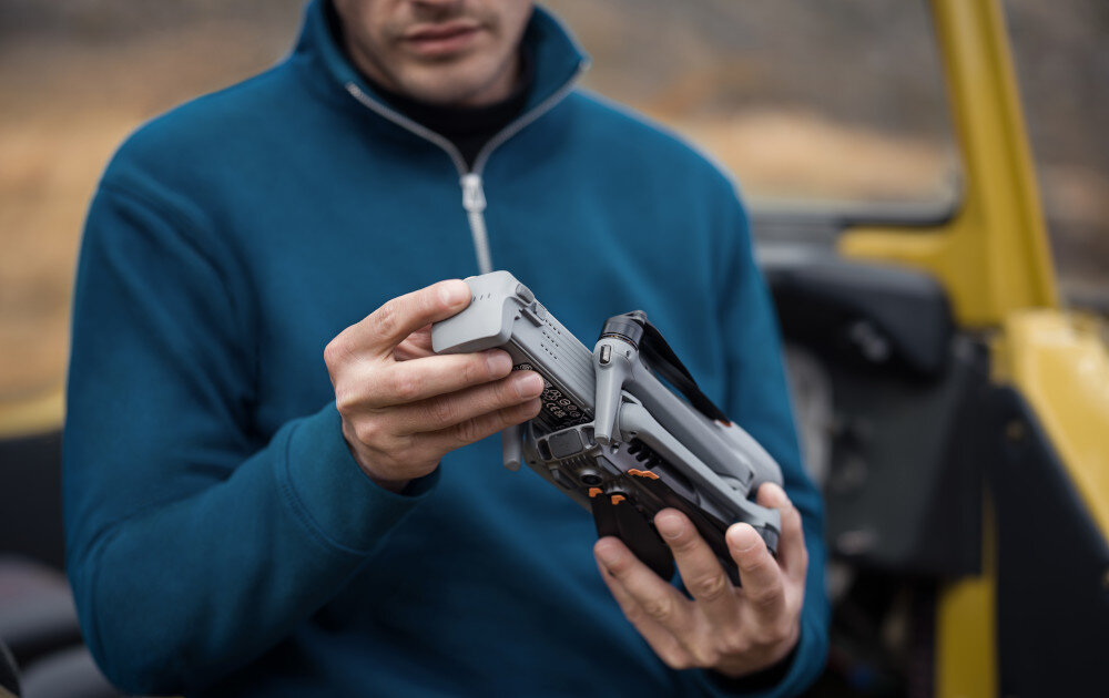 Osoba trzyma złożonego drona w rękach, ubrana w niebieską bluzę. W tle widoczny jest fragment żółtego pojazdu.