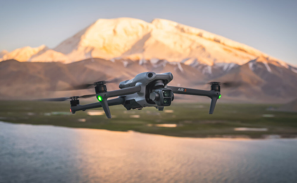Dron w locie nad wodą z widocznym ośnieżonym szczytem górskim w tle. Na ramieniu drona widoczny napis \'AIR 3\'.