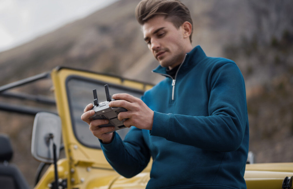 Osoba trzyma kontroler drona na tle żółtego pojazdu terenowego i górskiego krajobrazu.