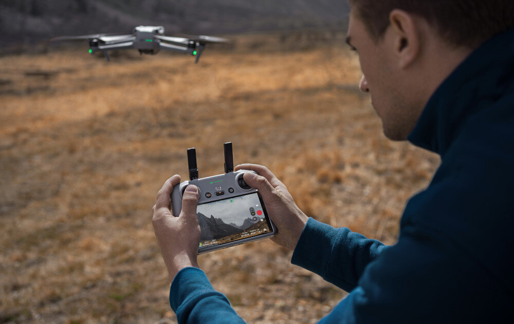 Osoba trzyma kontroler do drona, na ekranie którego widać krajobraz. W tle unosi się dron nad suchym terenem.
