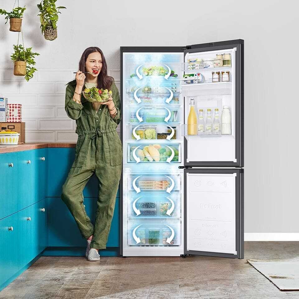 Kobieta stoi obok otwartej lodówki, trzymając miskę sałatki. Wnętrze lodówki jest wypełnione różnymi produktami spożywczymi, a na drzwiach widoczne są butelki i słoiki.