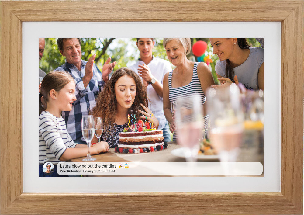 Ramka cyfrowa z drewnianą obwódką wyświetla zdjęcie grupy osób świętujących przy torcie. Na dole ekranu widoczny jest tekst: \'Laura blowing out the candles 
