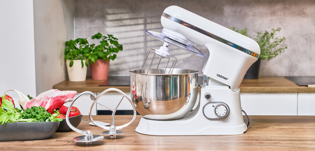 Na blacie kuchennym znajduje się biały robot planetarny z metalową misą i trzepaczką. Obok robota leżą dodatkowe mieszadła oraz miska z warzywami i owocami.