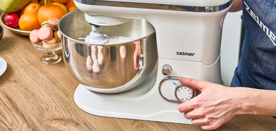 Na drewnianym blacie stoi biały robot planetarny z metalową misą, na której widnieje logo \'Zelmer\'. Obok robota znajduje się osoba, która trzyma pokrętło urządzenia.