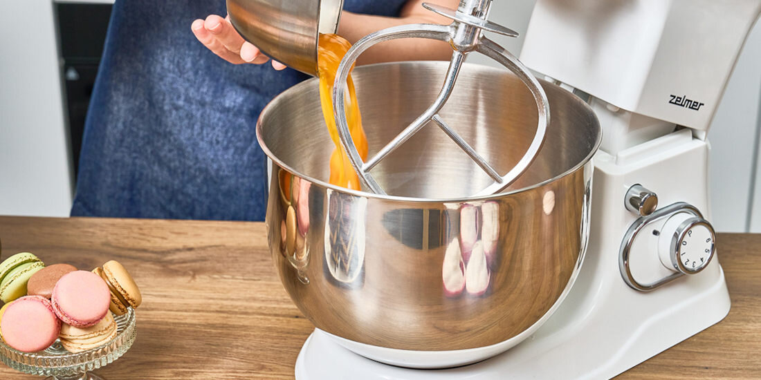Na blacie kuchennym stoi robot planetarny z metalową misą, w której znajduje się mieszadło. Obok na talerzyku leżą kolorowe makaroniki, a na urządzeniu widoczne jest logo \'Zelmer\'.