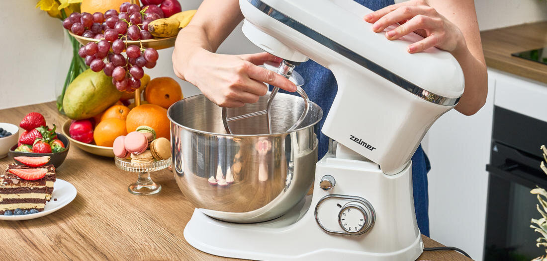 Osoba używa białego robota planetarnego marki Zelmer z metalową misą. W tle widoczne są owoce i ciasto na stole.