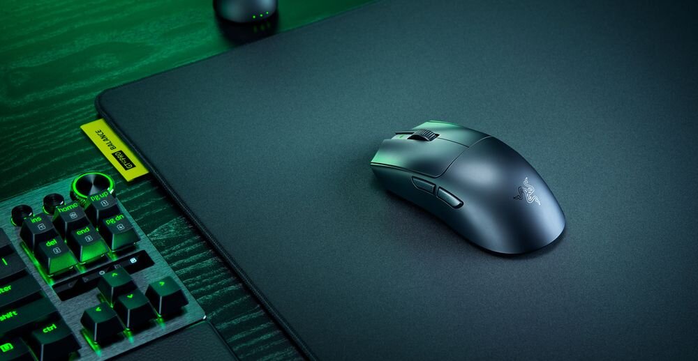 Na ciemnej podkładce znajduje się czarna mysz komputerowa z logo Razer. Obok widoczna jest część klawiatury z podświetlanymi klawiszami.