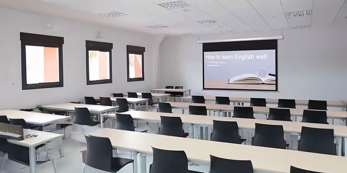 Sala lekcyjna z rzutnikiem wyświetlającym slajd z napisem \'How to learn English well\'. W pomieszczeniu znajdują się rzędy białych stołów i czarnych krzeseł.