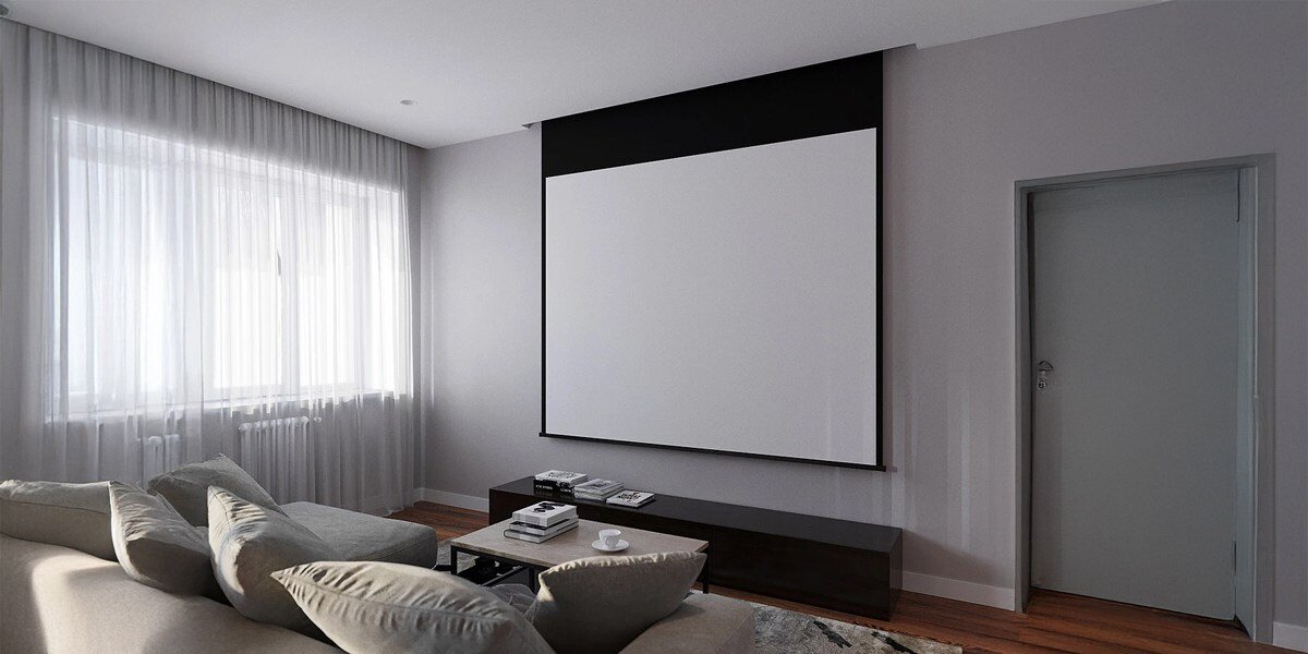 W salonie znajduje się duży ekran projekcyjny zamontowany na ścianie, obok którego stoi szara kanapa. Pomieszczenie jest jasne dzięki dużemu oknu z zasłonami.