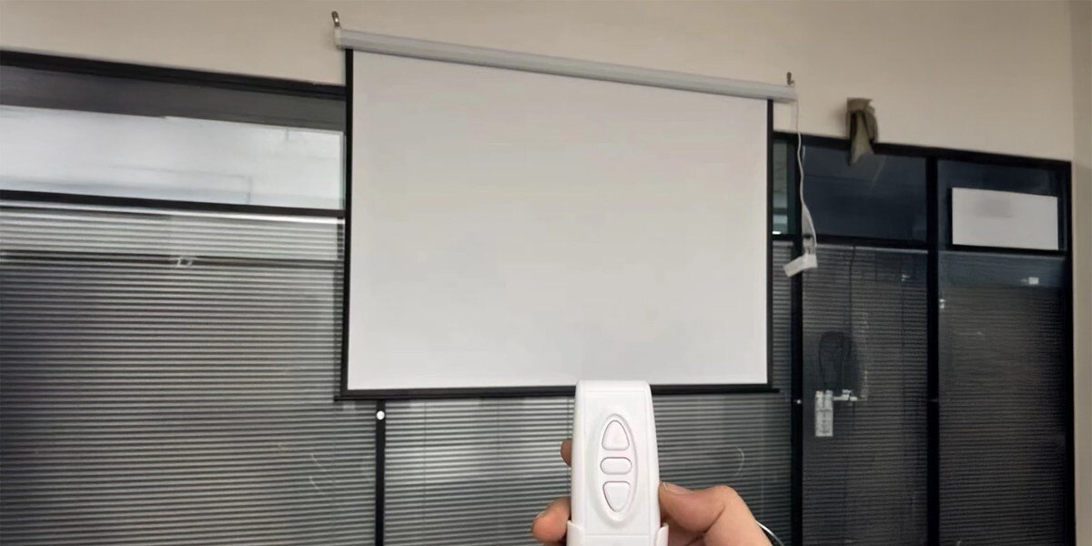 Na obrazie widoczny jest biały ekran projekcyjny zamontowany na ścianie oraz ręka trzymająca pilot z trzema przyciskami. W tle znajdują się przeszklone drzwi i okna z żaluzjami.