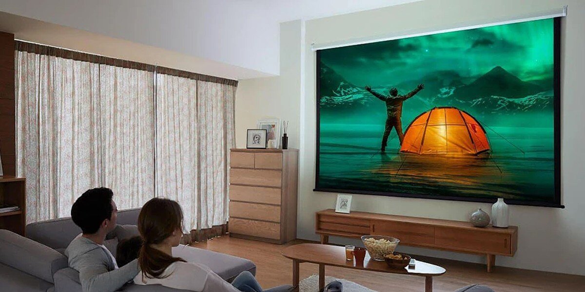 W salonie znajduje się ekran projekcyjny wyświetlający obraz osoby stojącej obok namiotu na tle gór i zorzy polarnej. Na kanapie siedzą trzy osoby oglądające projekcję.