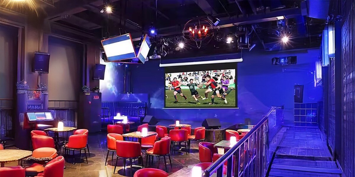 Wnętrze lokalu z czerwonymi krzesłami i okrągłymi stolikami, na ścianie wisi duży ekran projekcyjny wyświetlający mecz rugby. Pomieszczenie jest oświetlone niebieskimi światłami, a na suficie znajdują się reflektory i ekrany.