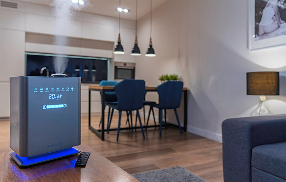 W nowoczesnym wnętrzu znajduje się nawilżacz ultradźwiękowy z wyświetlaczem pokazującym temperaturę 20.2°C i ikonami funkcji. W tle widać stół z krzesłami, lampy wiszące oraz fragment kuchni.