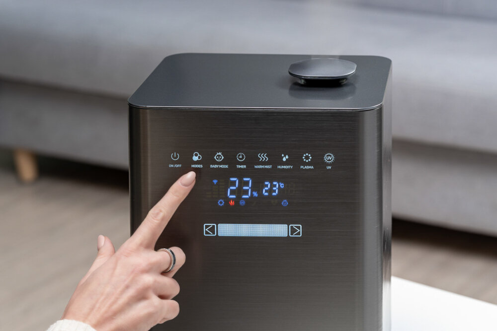 Osoba dotyka panelu sterowania nawilżacza ultradźwiękowego z wyświetlaczem pokazującym temperaturę 23°C. Na panelu widoczne są ikony: ON/OFF, MODES, BABY MODE, TIMER, MIST, HUMIDITY, PLASMA, UV.