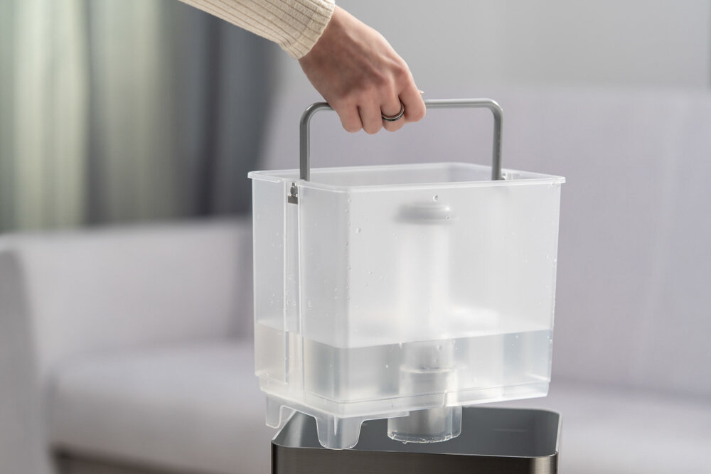 Osoba trzyma przezroczysty pojemnik z wodą za metalowy uchwyt. W tle widoczna jest rozmyta kanapa.