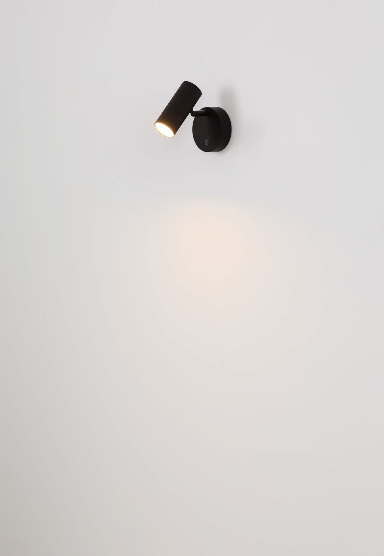 Na białej ścianie zamontowana jest czarna lampa reflektorowa, emitująca światło w dół. Lampa ma cylindryczny kształt i jest przymocowana do okrągłej podstawy.