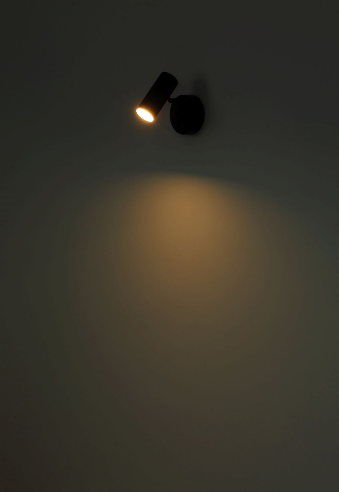 Na ciemnym tle widoczna jest włączona lampa reflektorowa, emitująca ciepłe światło na ścianę.