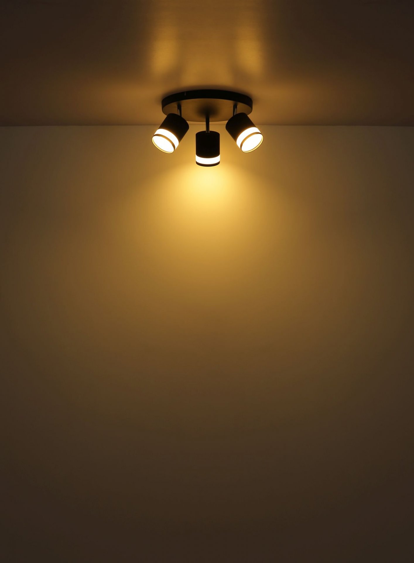 Na suficie zamontowana jest lampa sufitowa z trzema reflektorami, emitująca ciepłe, żółte światło. Światło oświetla ścianę poniżej, tworząc przytulną atmosferę.