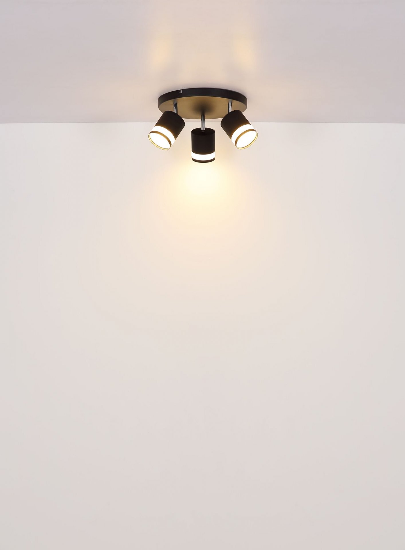 Na obrazie widoczna jest lampa sufitowa z trzema reflektorami, emitująca ciepłe światło. Lampa jest zamontowana na białym suficie.