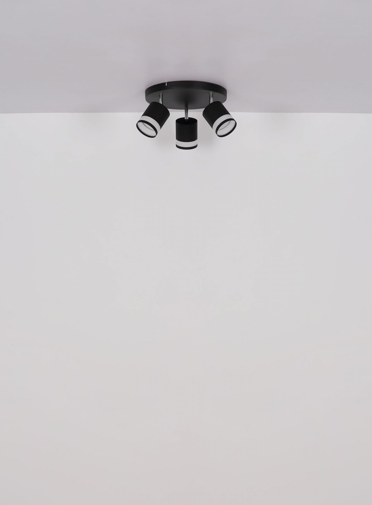 Na obrazie widoczna jest czarna lampa sufitowa z trzema reflektorami skierowanymi w różnych kierunkach. Lampa zamontowana jest na białym suficie.