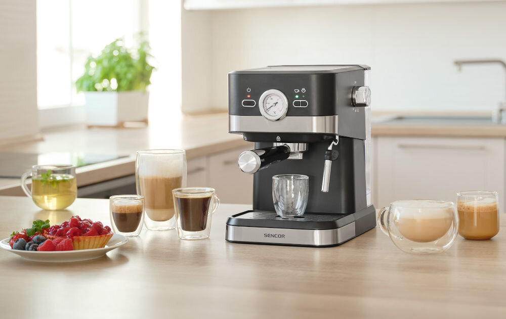 Na blacie kuchennym stoi czarny ekspres do kawy marki SENCOR z manometrem na przednim panelu. Obok ekspresu znajdują się różne filiżanki z napojami oraz talerz z owocami.
