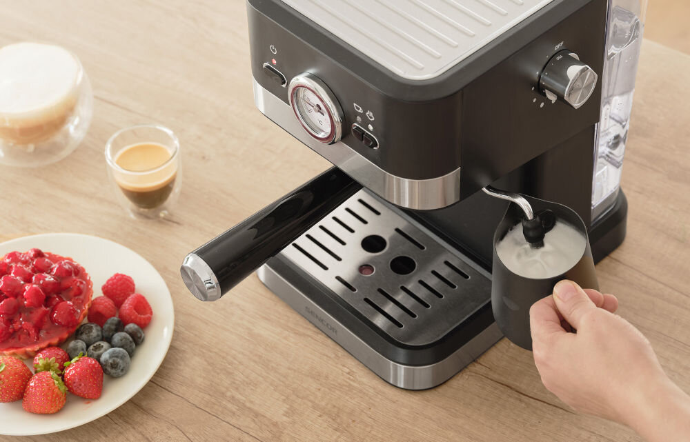 Czarny ekspres do kawy z metalicznymi elementami, na którym spieniane jest mleko w metalowym dzbanku. Obok znajduje się talerz z owocami i filiżanka z kawą.